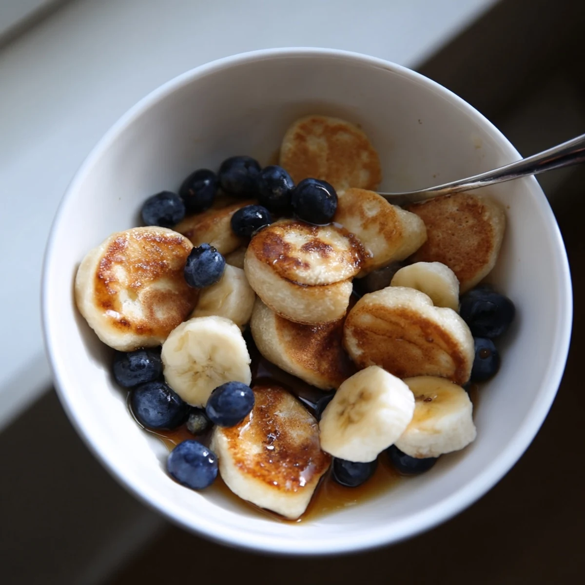 Golden mini pancake cereal stacked in a bowl, drizzled with maple syrup and cream.