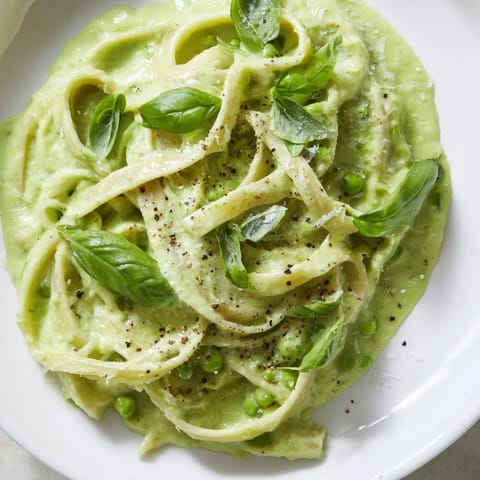 A close-up of silky Creamy Green Pea Alfredo sauce coating long pasta strands, topped with grated Parmesan and cracked black pepper.